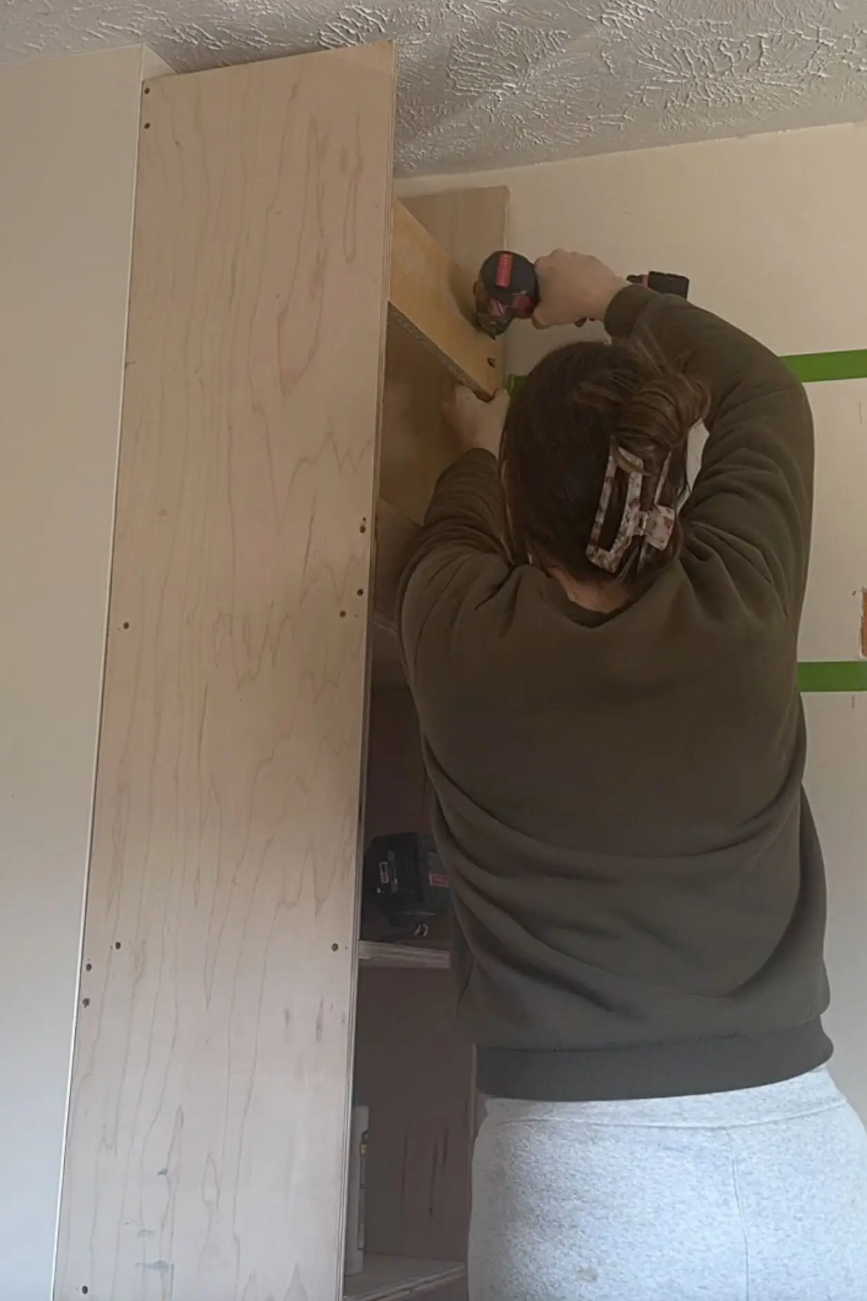 A woman drilling a wooden support beam into the side panel of a DIY built-in shelving unit. She is securing the upper structure, ensuring stability before adding additional shelves. Green painter’s tape marks the wall, indicating future design elements. The plywood panels are unfinished, showing the raw wood grain.